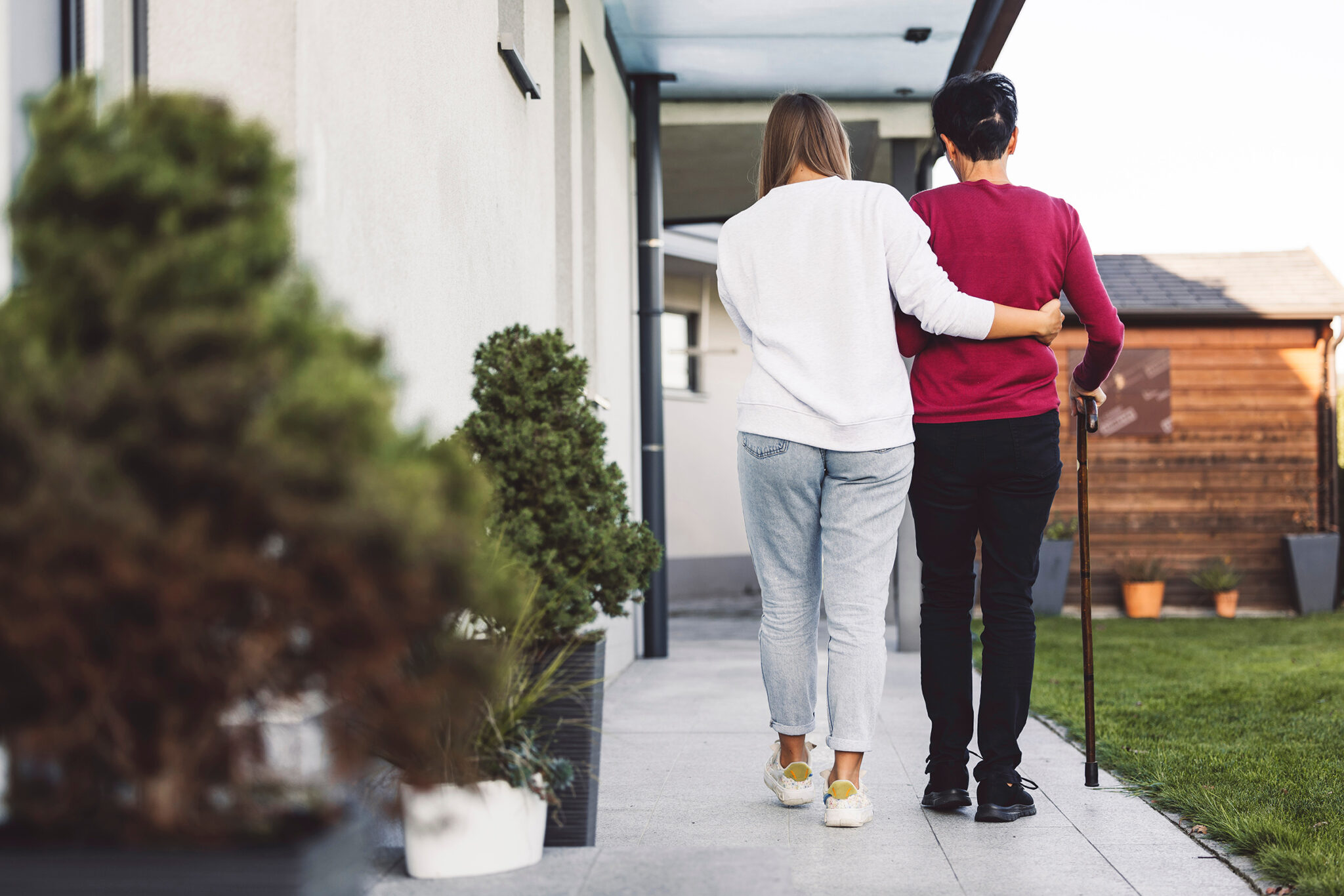 A female in a white shirt helping a senior lady in a red shirt and a cane walk who was assisted by the best senior care franchise. 